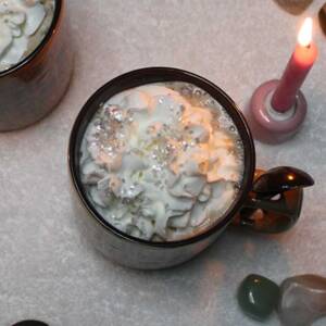 Two mugs of Love Potion #1: Winter's Embrace hot chocolate topped with whipped cream and edible silver flakes, surrounded by candles and crystals.