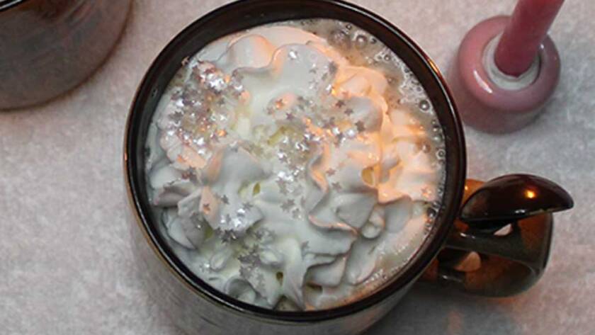 Two mugs of Love Potion #1: Winter's Embrace hot chocolate topped with whipped cream and edible silver flakes, surrounded by candles and crystals.