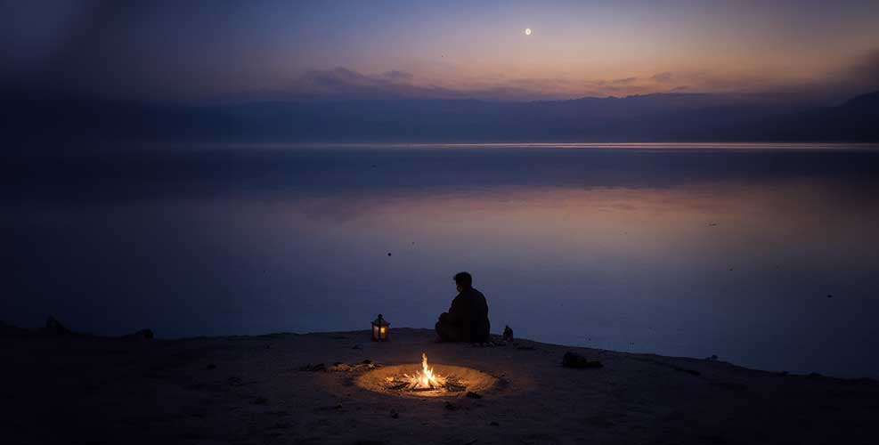 A solitary figure sits by a small campfire near a calm lake under a moonlit sky at dawn.