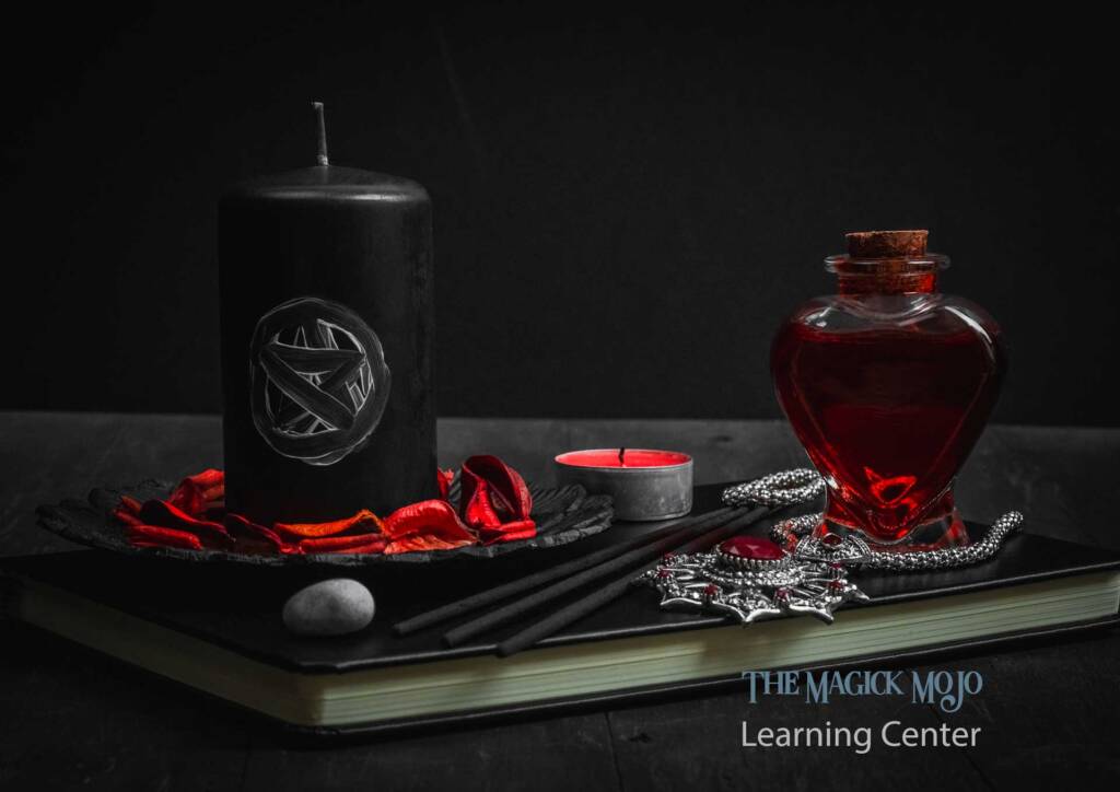 A bottle of Dove's Blood Ink next to a black candle with a pentagram, set on a dark surface with red petals and a book.