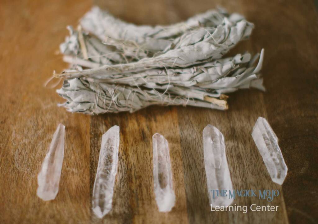 Five clear quartz crystals arranged in a row next to a bundle of white sage smudge sticks on a wooden table.