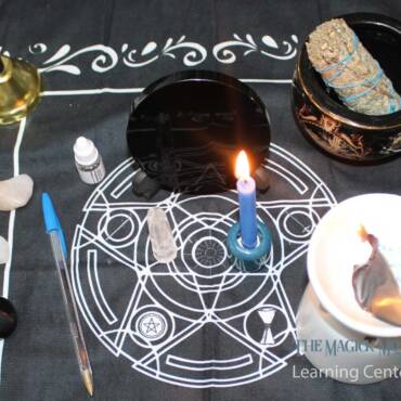 Setup for a Truth Revealing Spell featuring a blue candle, a mirror, clear quartz, a pen, sage, and other magical tools arranged on a mystical black cloth with white symbols.
