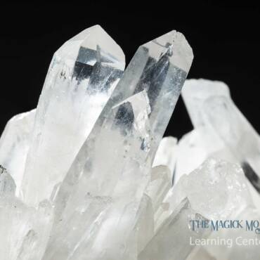 Close-up of a cluster of clear quartz crystals with sharp edges and transparent facets against a black background, branded with 'The Magick Mojo Learning Center'.