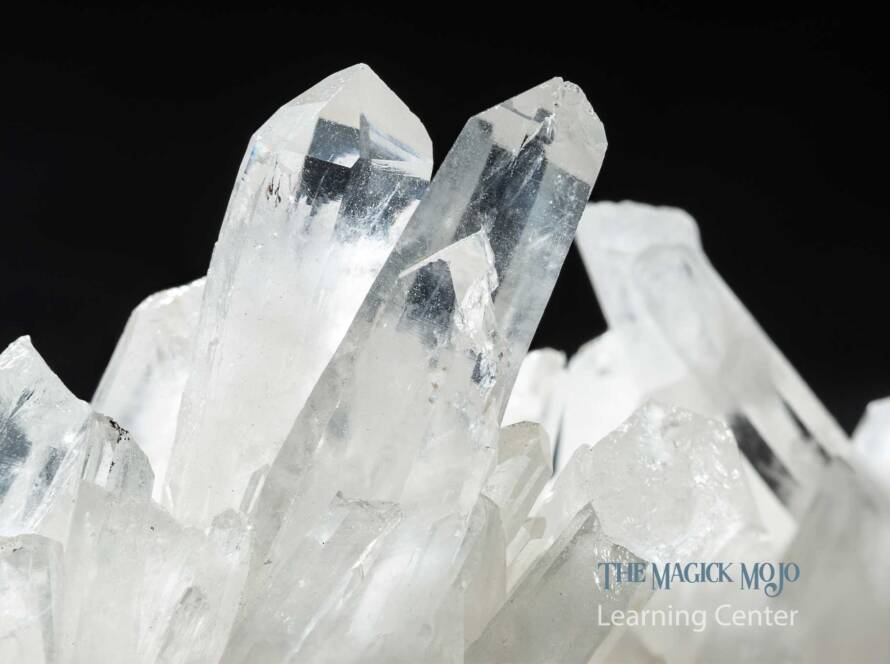 Close-up of a cluster of clear quartz crystals with sharp edges and transparent facets against a black background, branded with 'The Magick Mojo Learning Center'.