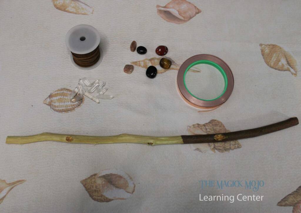 Materials for magic wand making including a stripped wood stick, assorted crystals, leather wrap, and copper tape on a seashell-patterned tablecloth.