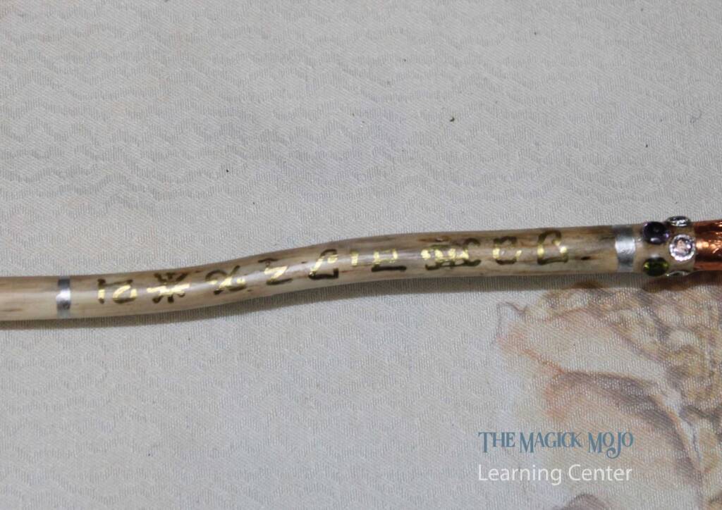 Close-up of a magic wand featuring golden inscriptions, embedded crystals, and copper wrapping on a textured background.