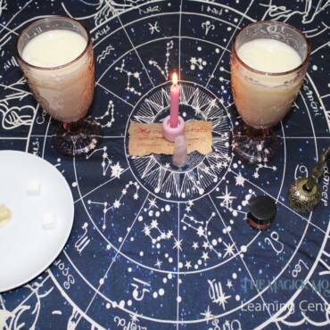 Two glasses of Love Potion #6, a pink candle, rose quartz crystal, spell paper, sugar cubes, and a quill on a celestial-themed tablecloth.