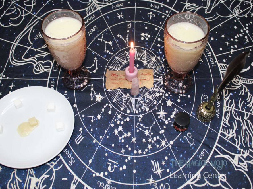 Two glasses of Love Potion #6, a pink candle, rose quartz crystal, spell paper, sugar cubes, and a quill on a celestial-themed tablecloth.